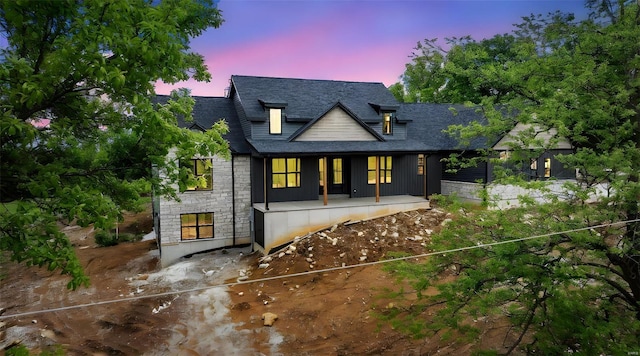 back of house at dusk with covered porch, a shingled roof, and stone siding