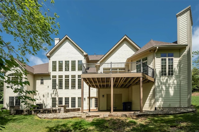 back of property with a shingled roof, a patio area, a lawn, a chimney, and a deck