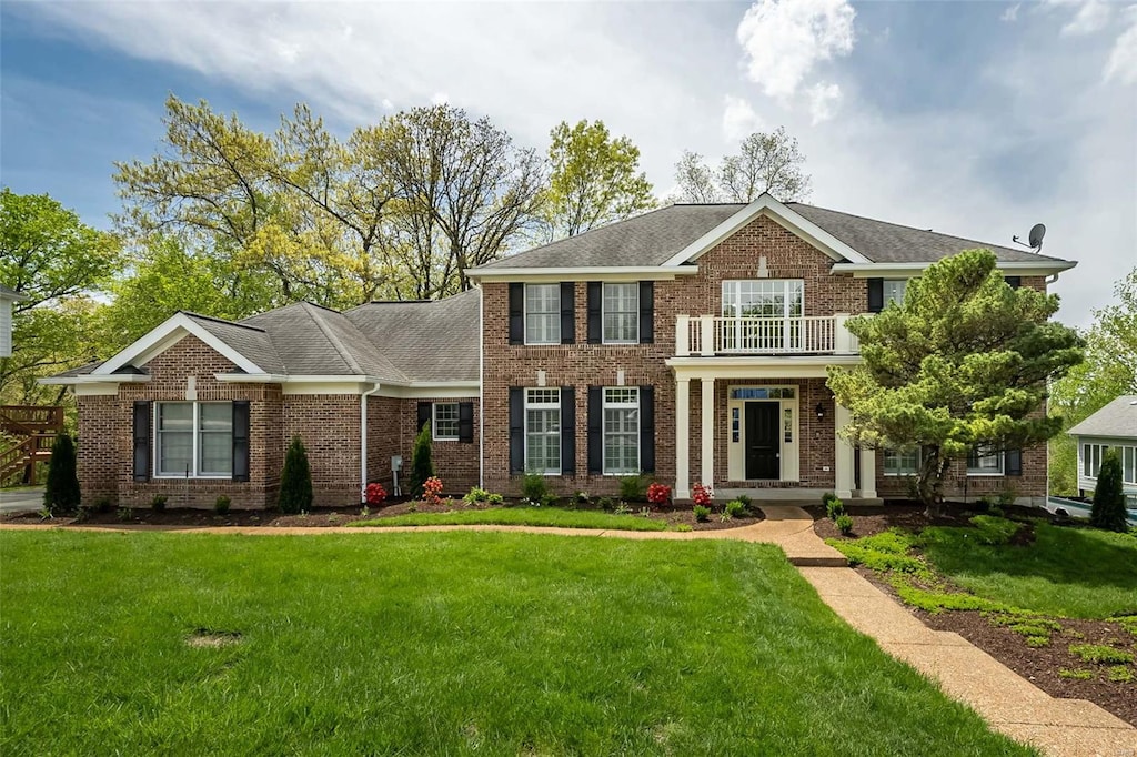 colonial home featuring a front lawn, a balcony, a shingled roof, and brick siding