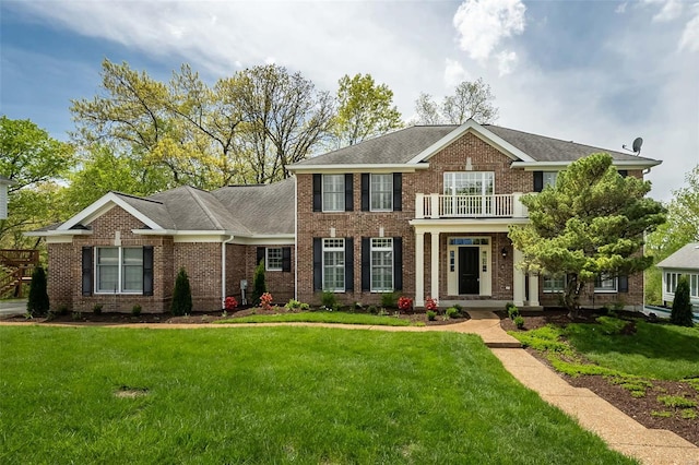colonial home featuring a front lawn, a balcony, a shingled roof, and brick siding