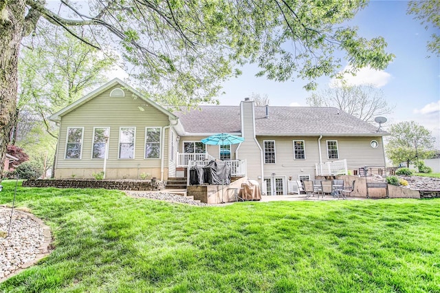 rear view of property with a patio, a deck, a chimney, a shingled roof, and a lawn