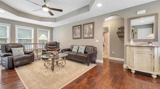 living area with ceiling fan, a raised ceiling, baseboards, arched walkways, and dark wood finished floors