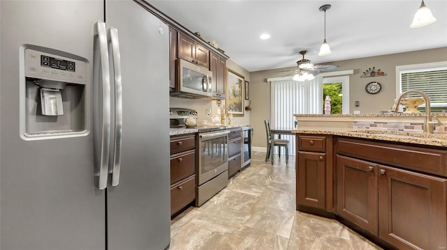 kitchen with light stone counters, a sink, stainless steel appliances, decorative light fixtures, and a ceiling fan