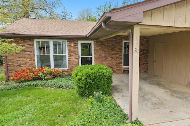 view of exterior entry featuring brick siding, a shingled roof, a yard, and an attached carport