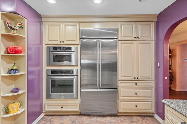 kitchen with cream cabinets, light stone counters, built in features, arched walkways, and appliances with stainless steel finishes