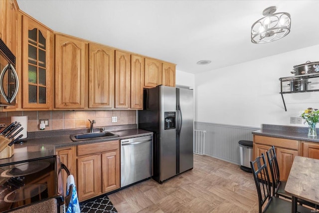 kitchen with appliances with stainless steel finishes, dark countertops, a sink, a wainscoted wall, and glass insert cabinets
