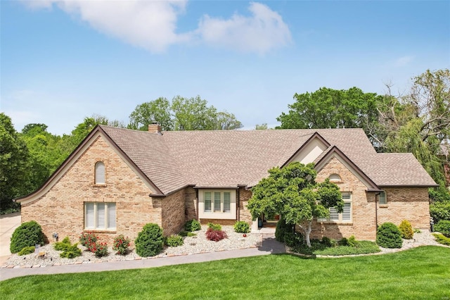 view of front of property featuring a shingled roof, a front lawn, a chimney, and brick siding