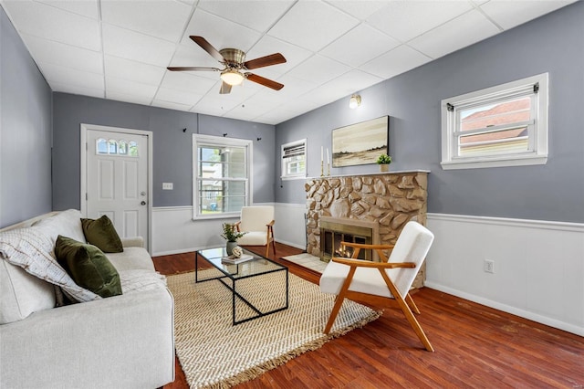 living area featuring a stone fireplace, a ceiling fan, a drop ceiling, and wood finished floors