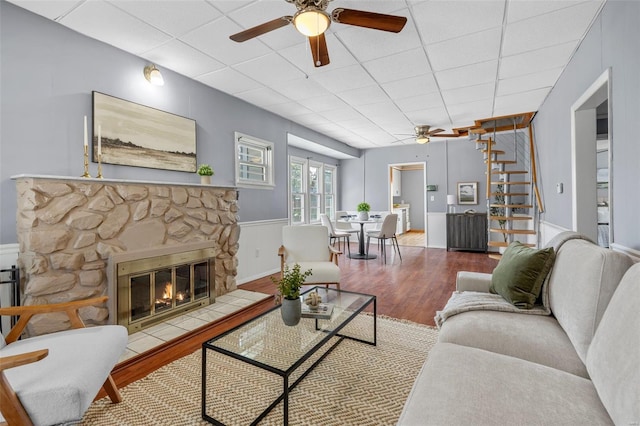 living room featuring stairs, wood finished floors, a stone fireplace, a paneled ceiling, and a ceiling fan