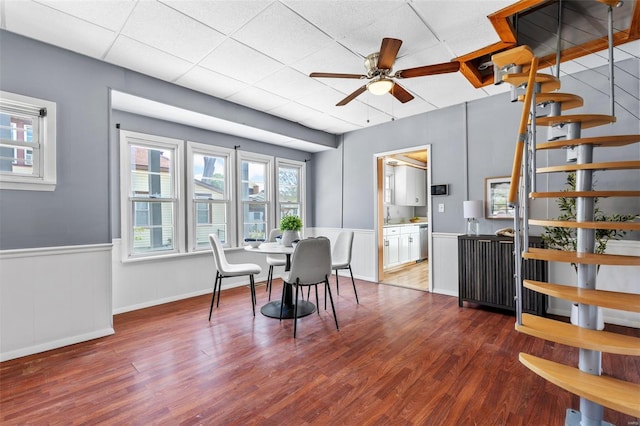 dining room with a ceiling fan, a drop ceiling, wood finished floors, and a wainscoted wall