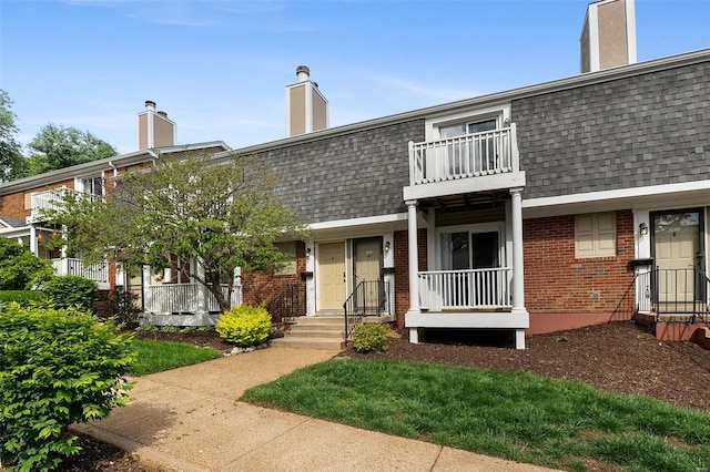 view of front facade with a shingled roof, a chimney, brick siding, and a balcony