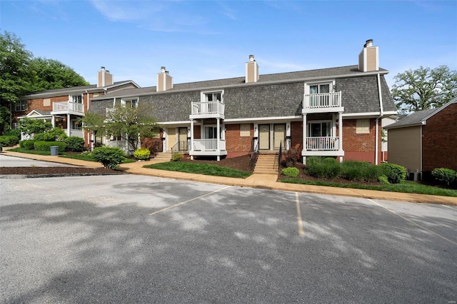 view of front of property featuring brick siding, a chimney, a balcony, mansard roof, and uncovered parking