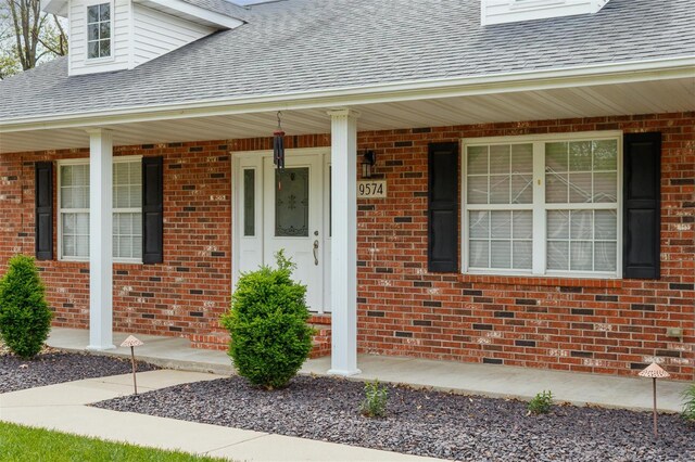 doorway to property with a shingled roof, a porch, and brick siding