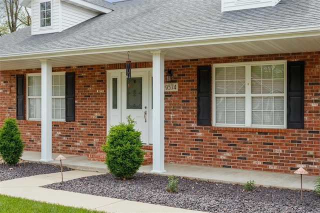 doorway to property with a shingled roof, a porch, and brick siding