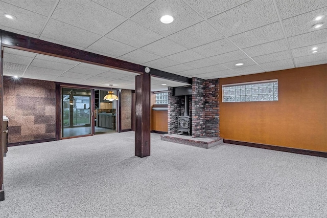 basement featuring baseboards, a wood stove, a paneled ceiling, and carpet