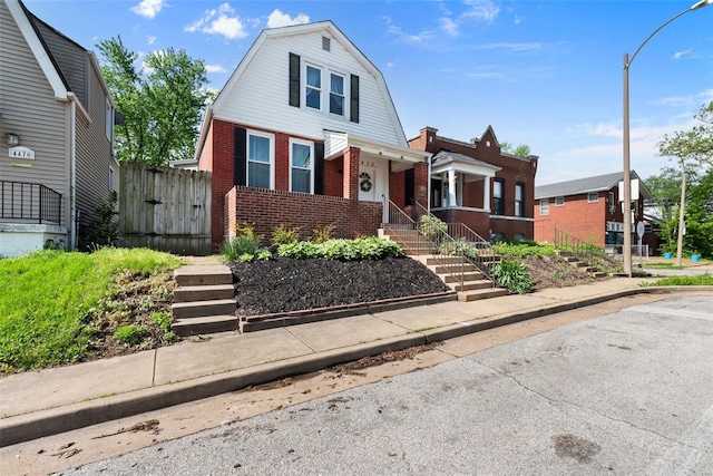 dutch colonial with covered porch, brick siding, fence, a gambrel roof, and stairs