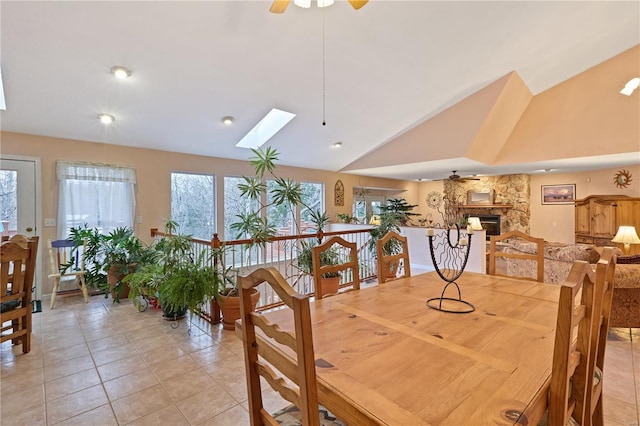 dining area featuring a stone fireplace, a ceiling fan, lofted ceiling with skylight, and light tile patterned floors