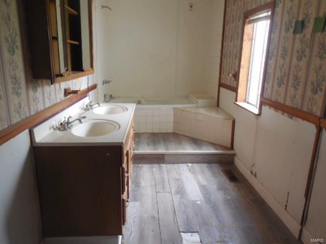 bathroom with tiled bath, wallpapered walls, a sink, and wood finished floors