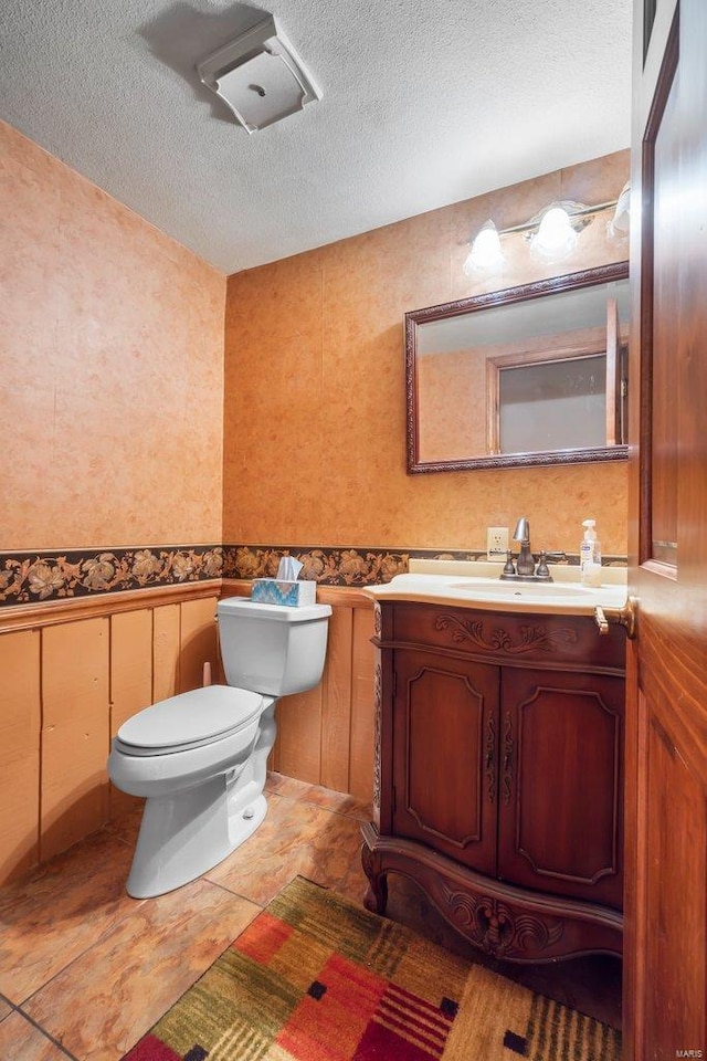 half bath with wainscoting, vanity, toilet, a textured ceiling, and wallpapered walls