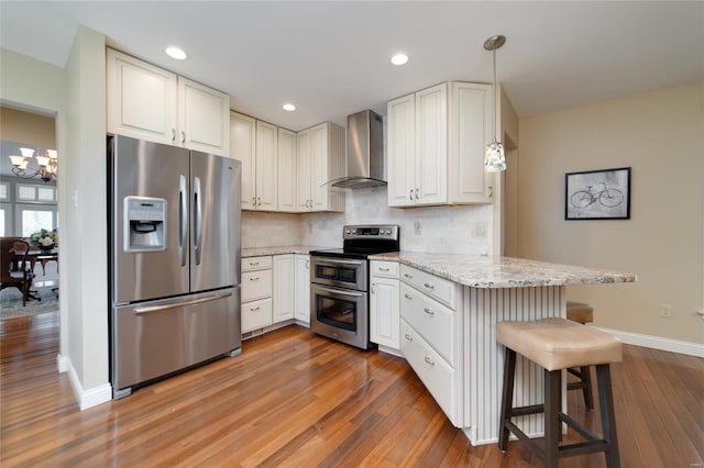 kitchen with a peninsula, baseboards, wall chimney exhaust hood, a chandelier, and appliances with stainless steel finishes