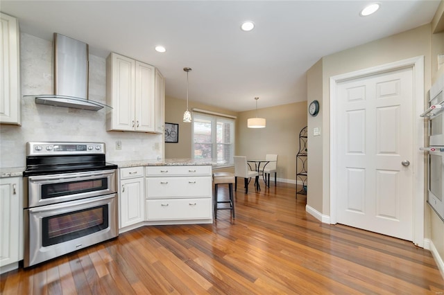kitchen featuring wall chimney range hood, white cabinets, hanging light fixtures, light wood-style floors, and range with two ovens