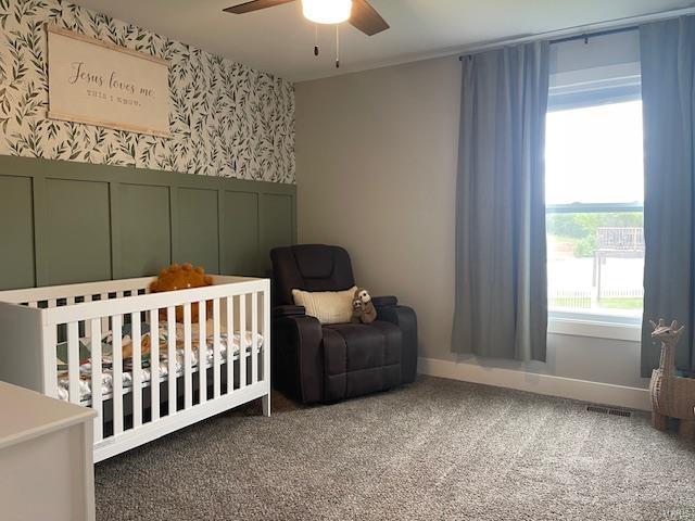 carpeted bedroom featuring wallpapered walls, wainscoting, ceiling fan, visible vents, and a crib