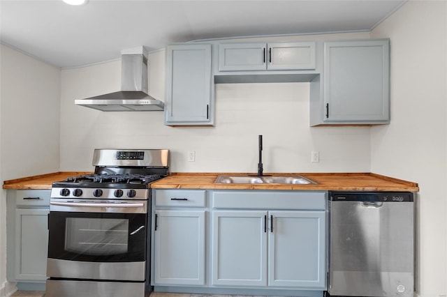 kitchen with butcher block countertops, wall chimney exhaust hood, gray cabinets, a sink, and stainless steel appliances