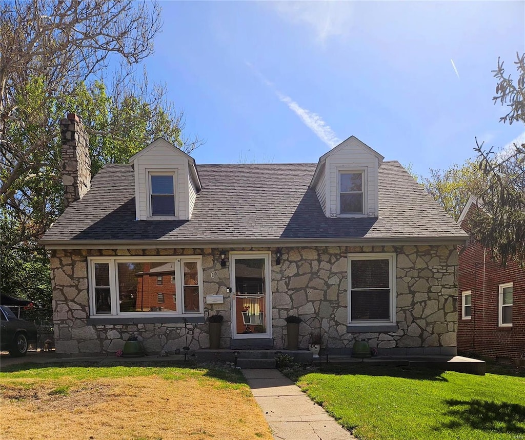 cape cod home featuring stone siding, roof with shingles, a chimney, and a front lawn