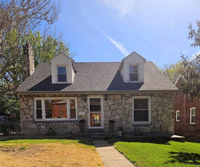 cape cod home featuring stone siding, roof with shingles, a chimney, and a front lawn