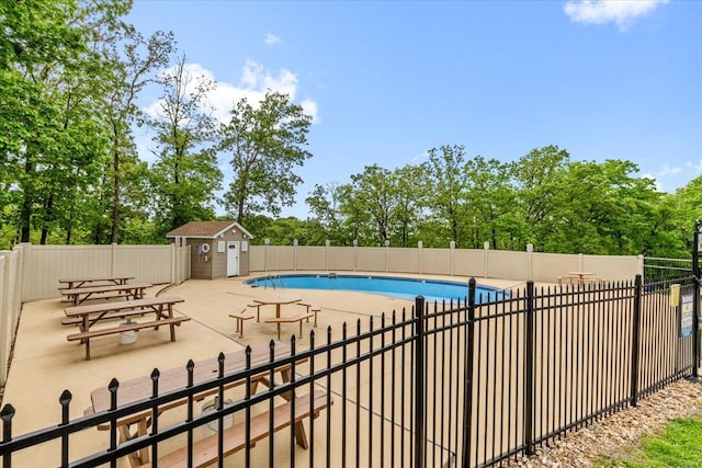 community pool featuring fence, a patio, an outdoor structure, and view of wooded area