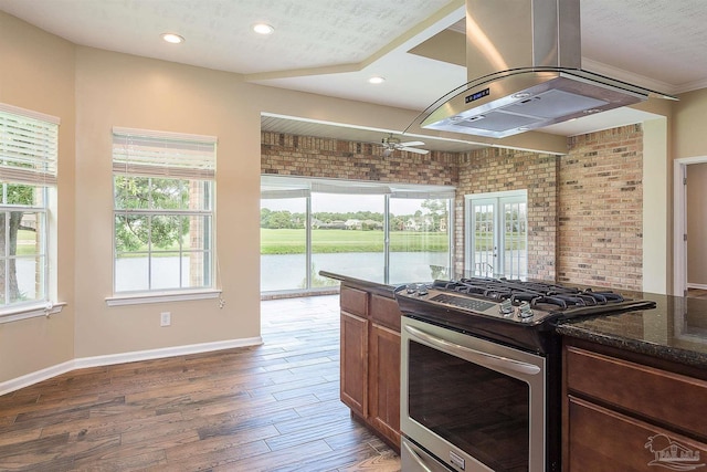 kitchen with stainless steel gas stove, dark stone countertops, dark hardwood / wood-style floors, a wealth of natural light, and island exhaust hood