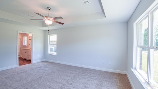 unfurnished bedroom featuring multiple windows, connected bathroom, ceiling fan, and a tray ceiling