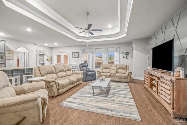 living area with arched walkways, a ceiling fan, light wood-type flooring, a raised ceiling, and crown molding