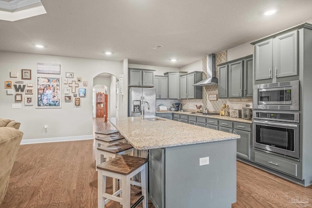 kitchen featuring arched walkways, appliances with stainless steel finishes, gray cabinetry, wall chimney range hood, and a sink