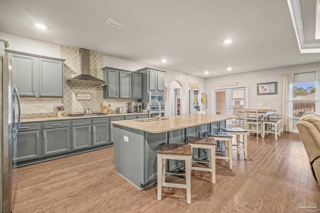 kitchen with arched walkways, stainless steel appliances, a kitchen island with sink, wall chimney range hood, and a kitchen bar