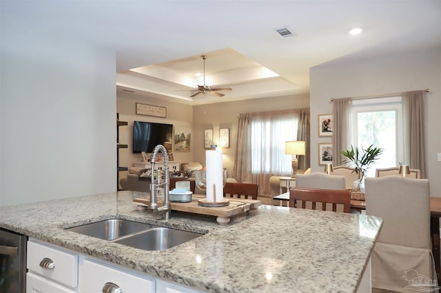 kitchen featuring white cabinets, sink, ceiling fan, a tray ceiling, and light stone counters