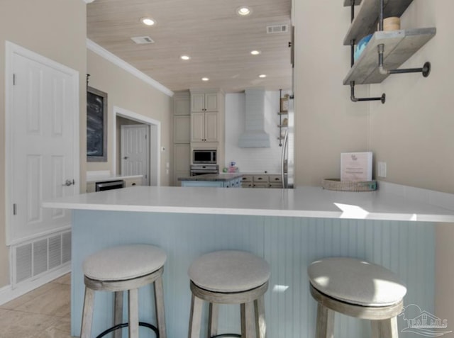 kitchen with wall chimney range hood, crown molding, a breakfast bar, stainless steel microwave, and kitchen peninsula