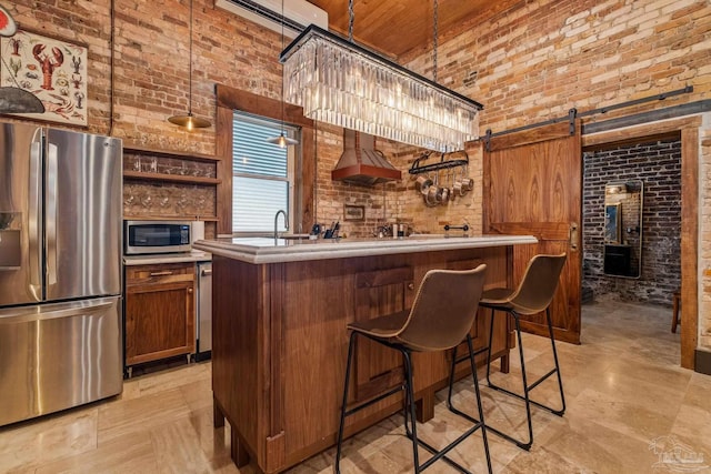 bar with appliances with stainless steel finishes, brick wall, wall chimney range hood, a barn door, and hanging light fixtures