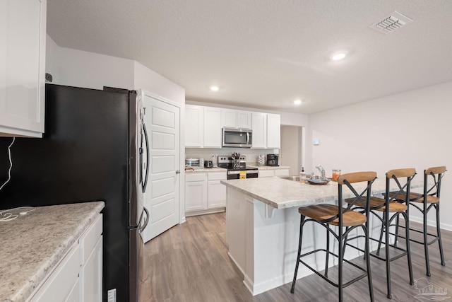 kitchen with a kitchen bar, stainless steel appliances, white cabinets, light hardwood / wood-style floors, and an island with sink