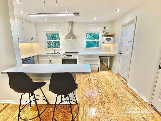 kitchen featuring light wood-type flooring, sink, white cabinetry, wall chimney range hood, and stainless steel appliances