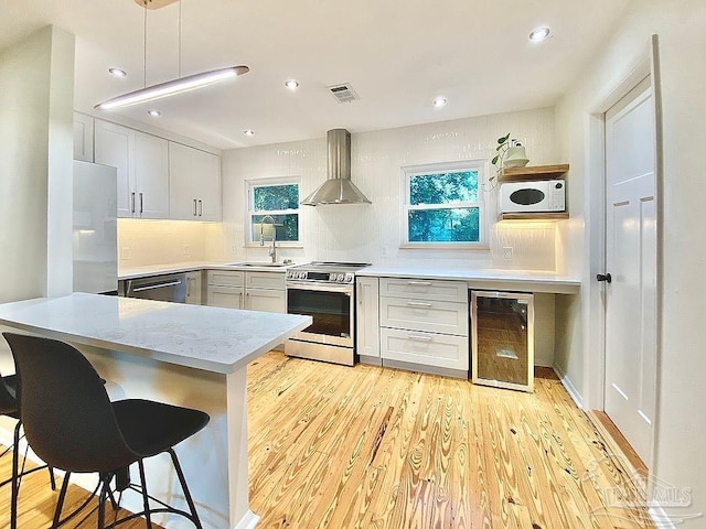 kitchen featuring appliances with stainless steel finishes, light hardwood / wood-style floors, beverage cooler, decorative light fixtures, and wall chimney range hood