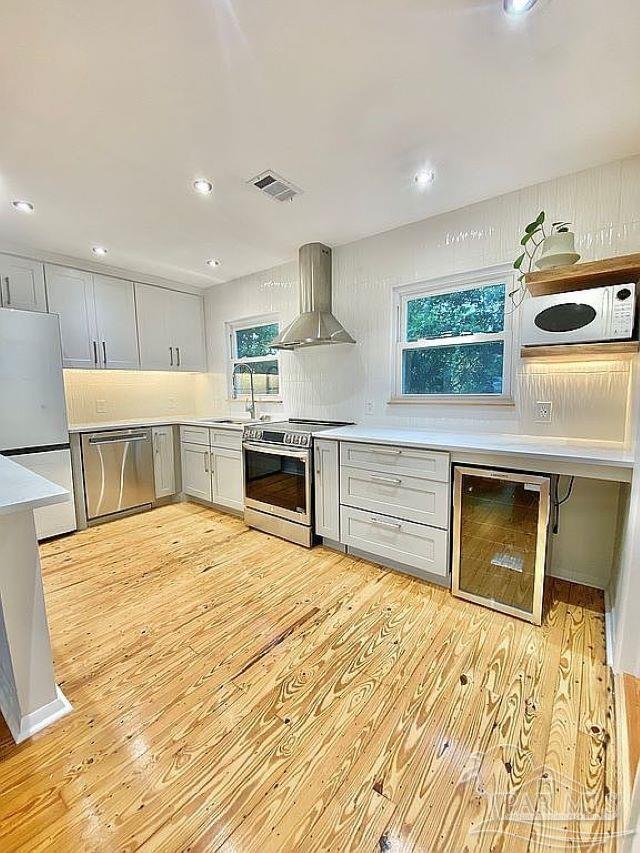 kitchen with light wood-type flooring, tasteful backsplash, beverage cooler, wall chimney range hood, and appliances with stainless steel finishes