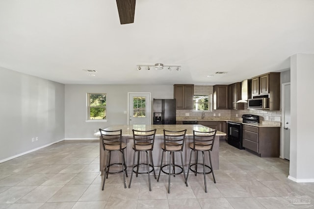 kitchen featuring black appliances, wall chimney exhaust hood, a kitchen island, and a healthy amount of sunlight