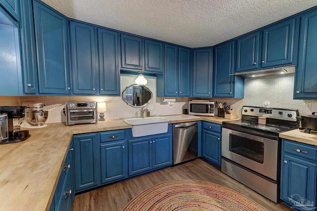 kitchen featuring blue cabinetry, appliances with stainless steel finishes, a sink, under cabinet range hood, and dark wood-type flooring