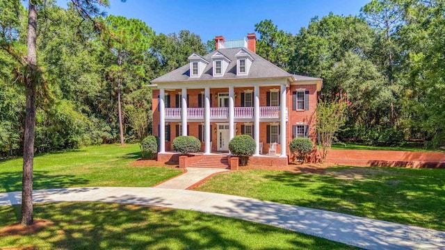 greek revival inspired property featuring a chimney, a porch, a front yard, a balcony, and brick siding
