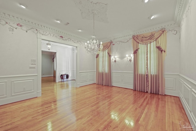 empty room with a decorative wall, light wood-type flooring, a chandelier, crown molding, and wainscoting