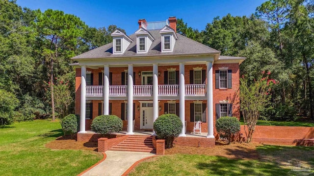 greek revival inspired property with covered porch, brick siding, a chimney, and a balcony