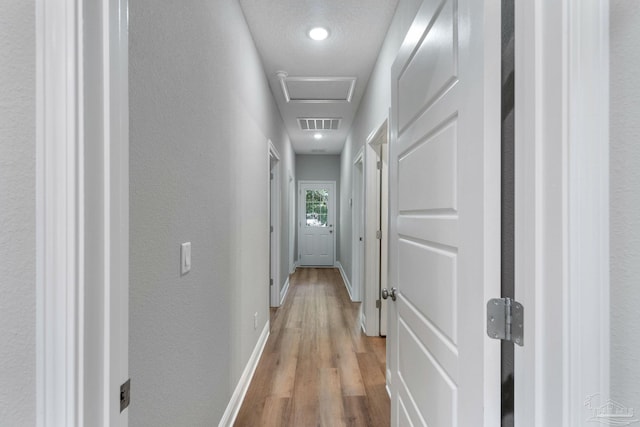 hall featuring light wood-style flooring, attic access, baseboards, and a textured wall