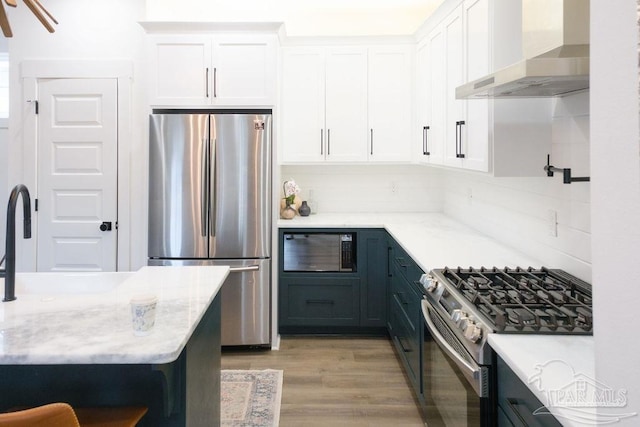 kitchen featuring light stone countertops, appliances with stainless steel finishes, light wood-style floors, white cabinetry, and wall chimney exhaust hood