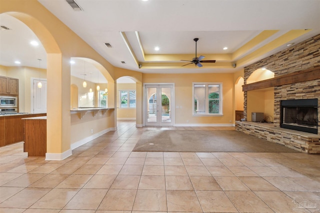 unfurnished living room featuring french doors, a stone fireplace, a tray ceiling, light tile patterned floors, and ceiling fan